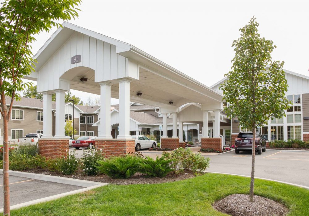 Drive-through portico with white pillars and brick accents at building entrance, surrounded by greenery and parked cars on a clear day.