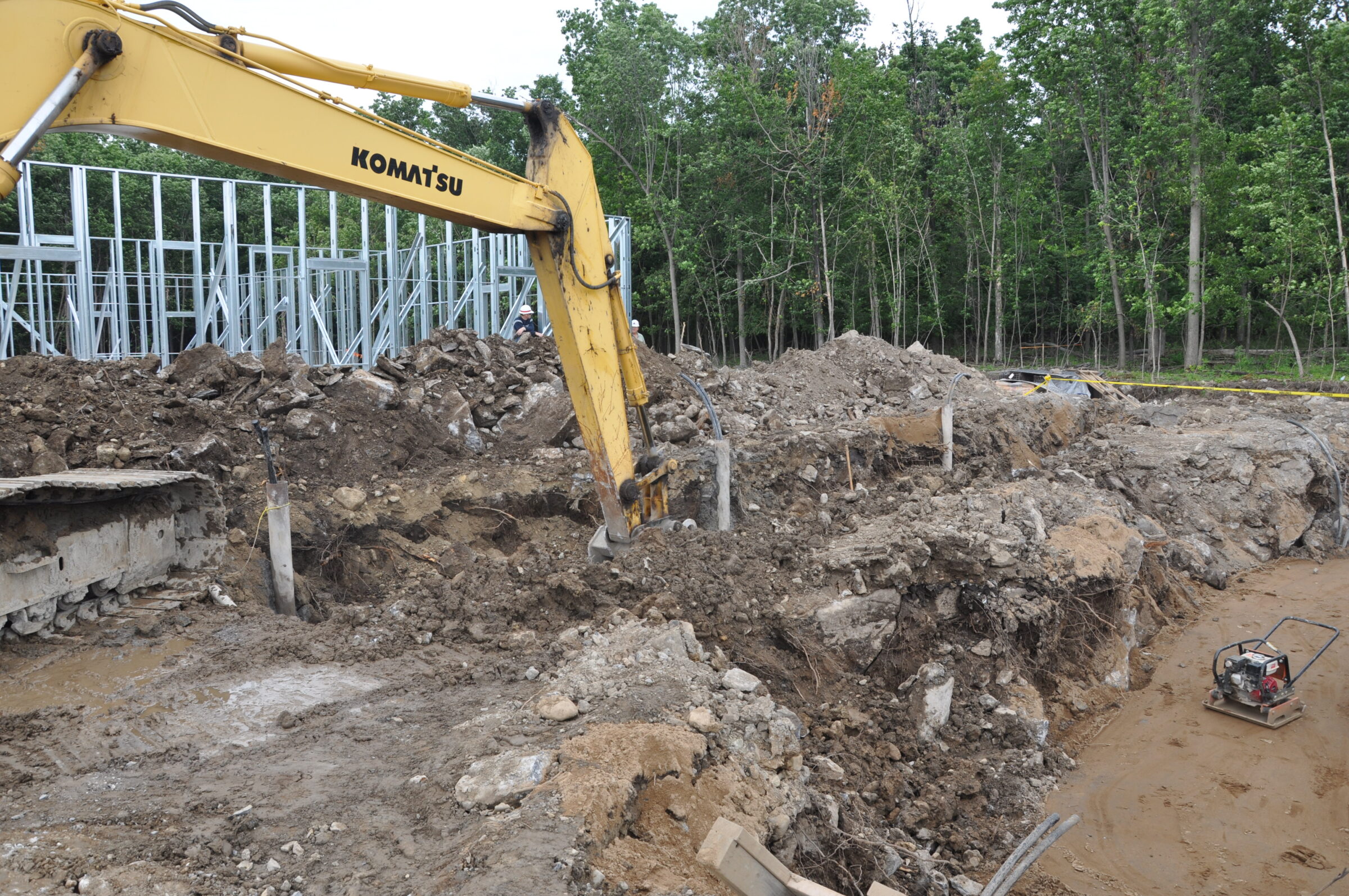 A construction site with a Komatsu excavator digging earth near a steel frame structure, surrounded by trees and machinery.